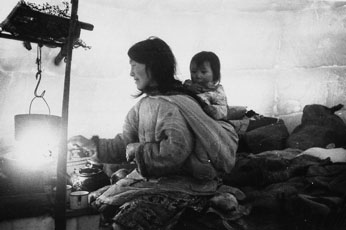 À l'intérieur d'un igloo, une femme ravive le qulliq, elle porte son enfant dans son amautik (Salluit, vers 1950)